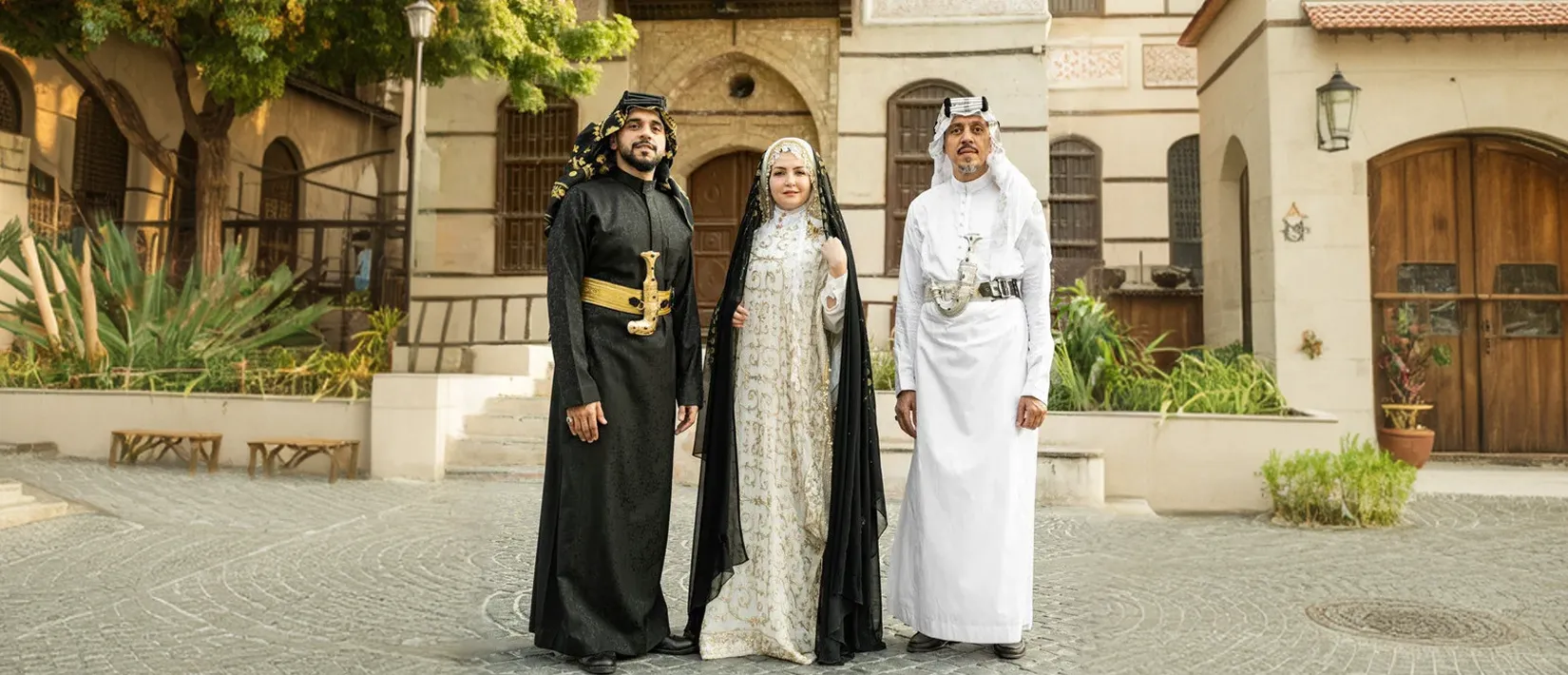 Three individuals dressed in traditional Saudi Arabian cultural clothing standing in front of a historic building.
