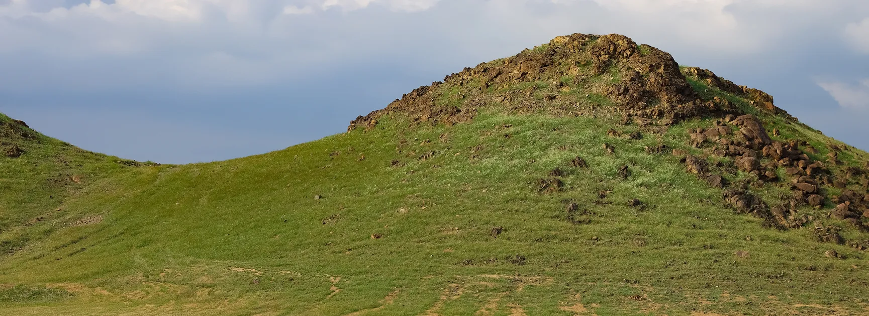 A scenic view of green grassy hills with rocky terrain under a cloudy sky in Makkah.