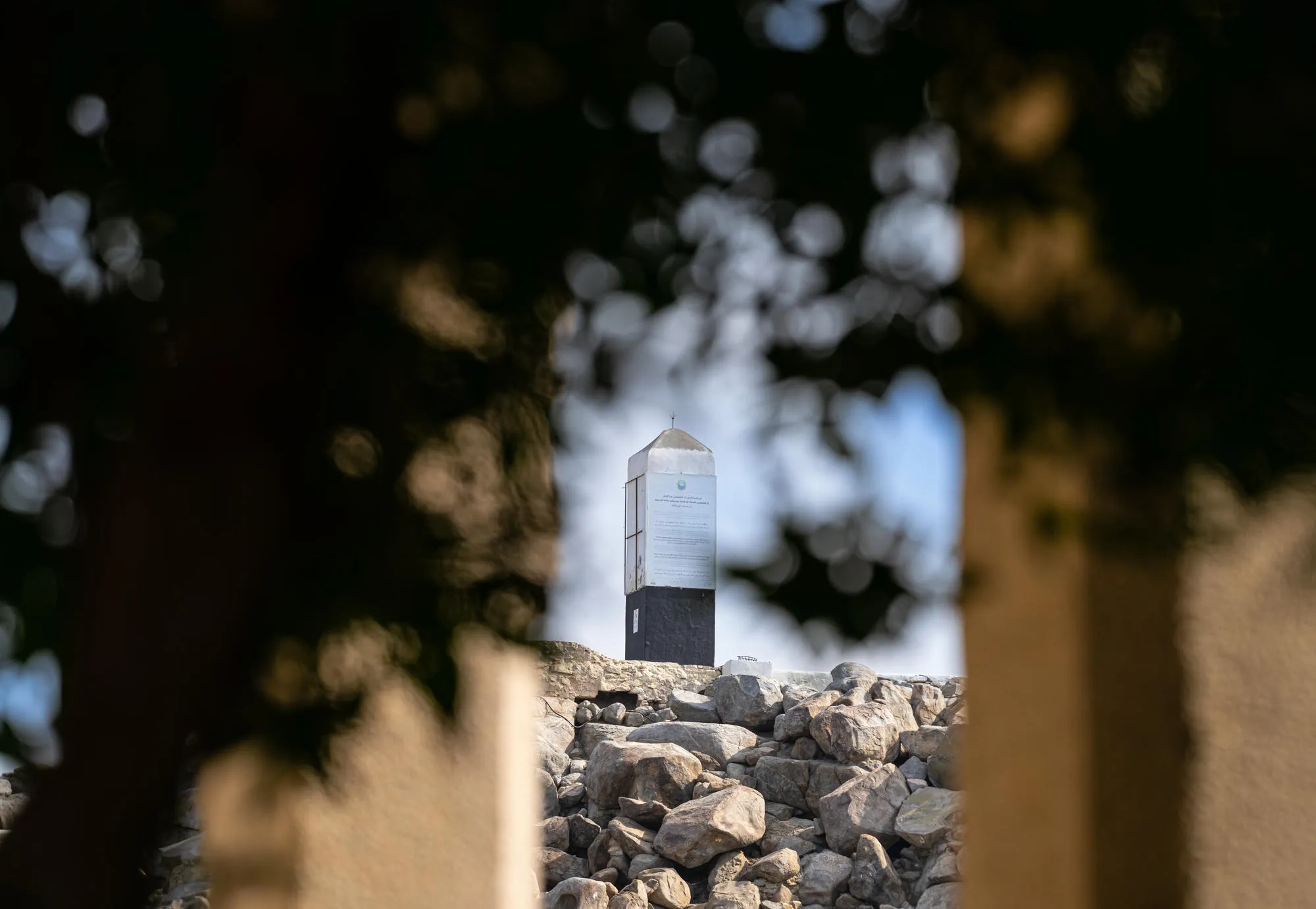 A framed view through tree branches of the white landmark pillar at the top of Jabal al-Rahmah (Mountain of Mercy) in Makkah.